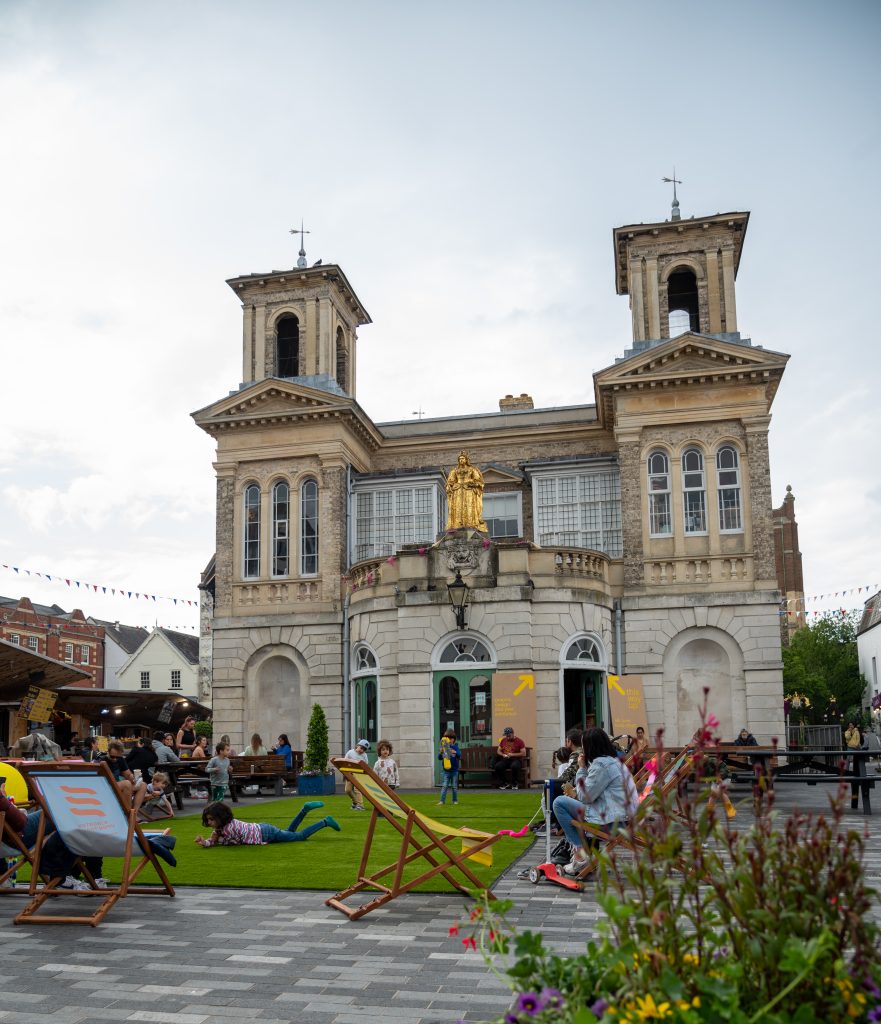 Image showing brick building, with gold statue in the background, with plants and deck chairs in the foreground.