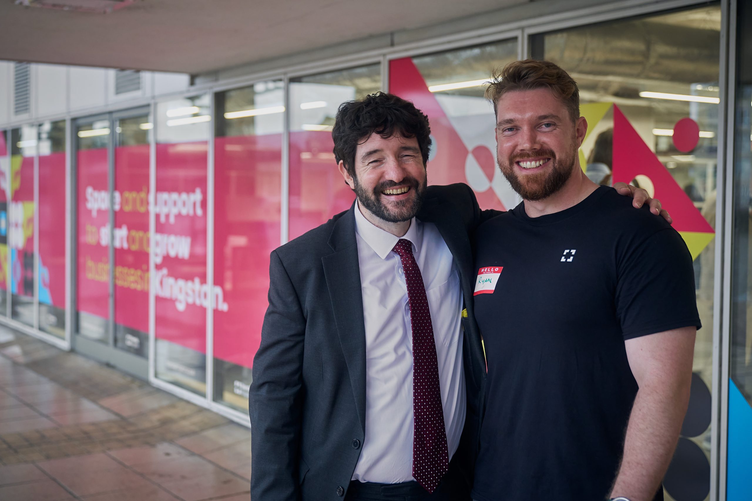 Kingston Council Leader, Andreas Kirsch with Town Square Kingston Hub Manager Ryan Gough Image shows Kingston Council Leader Andreas Kirsch, wearing a black suit, white short and red spotted tie, with Town Square Kingston Hub Manager Ryan Gough wearing a black t-shirt, both in front of the glass panelled doors of an office building.